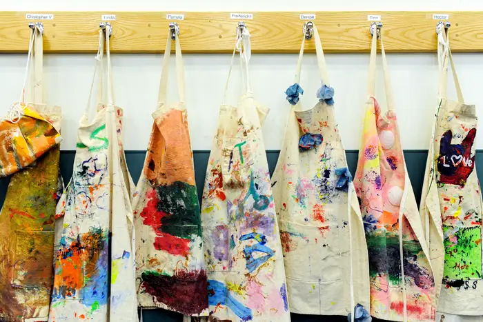 Aprons, covered in colorful paint, hang on hooks on a wall, with names next to the hooks